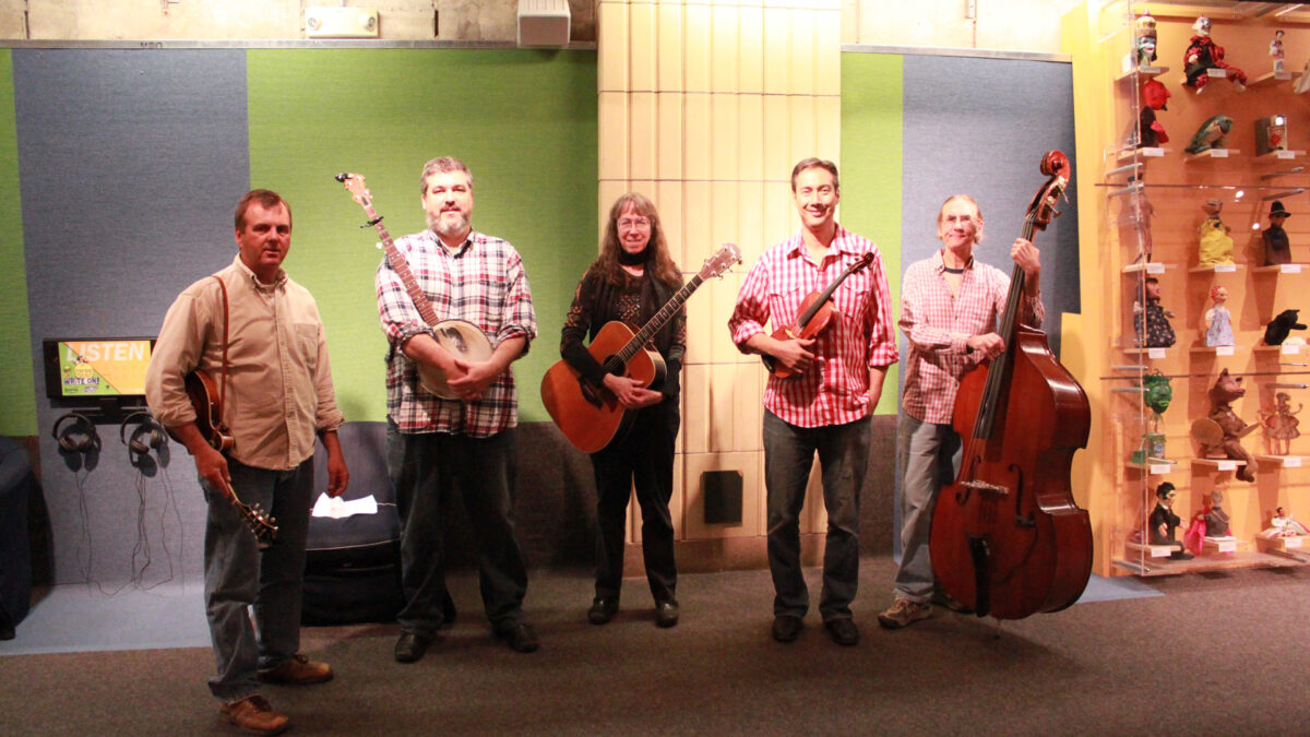 An adult holding a mandolin, an adult holding a banjo, an adult holding an acoustic guitar, an adult holding a violin, and an adult holding an upright bass, all standing in profile