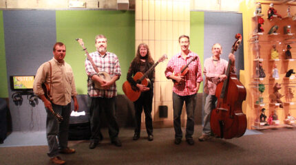 An adult holding a mandolin, an adult holding a banjo, an adult holding an acoustic guitar, an adult holding a violin, and an adult holding an upright bass, all standing in profile