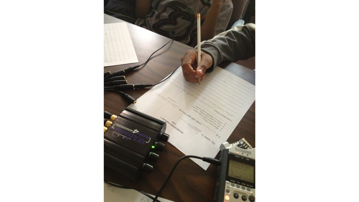 photo-1-(6)_wp A youth writing on a piece of paper sitting behind a headphone splitter and a portable recorder on a brown table