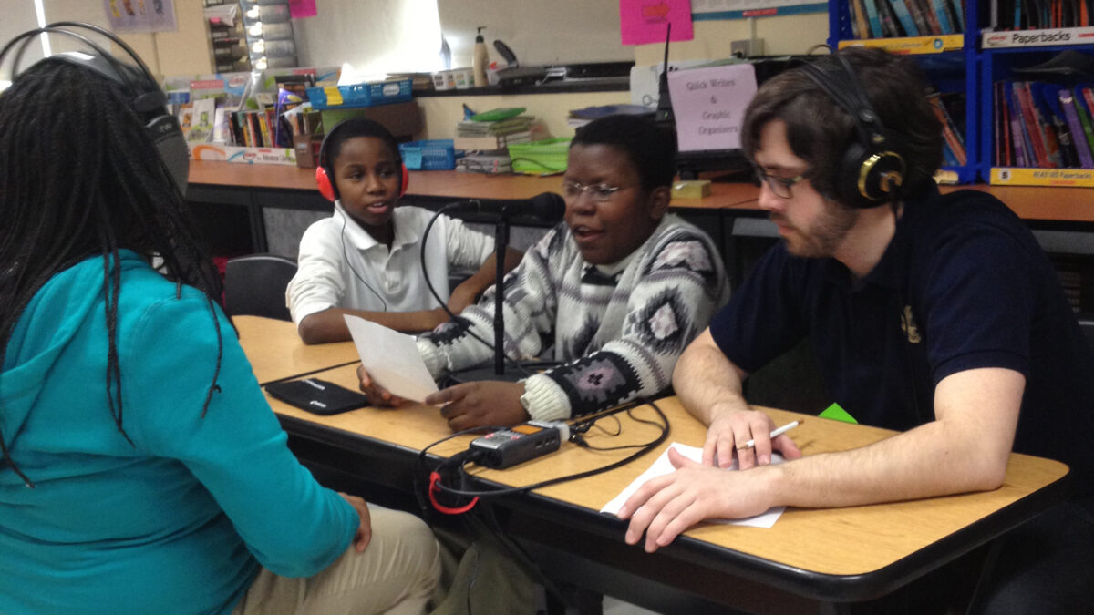 Two youth and an adult wearing headphones listening to a youth speaking into a microphone connected to a portable recorder and reading a piece of paper in a classroom