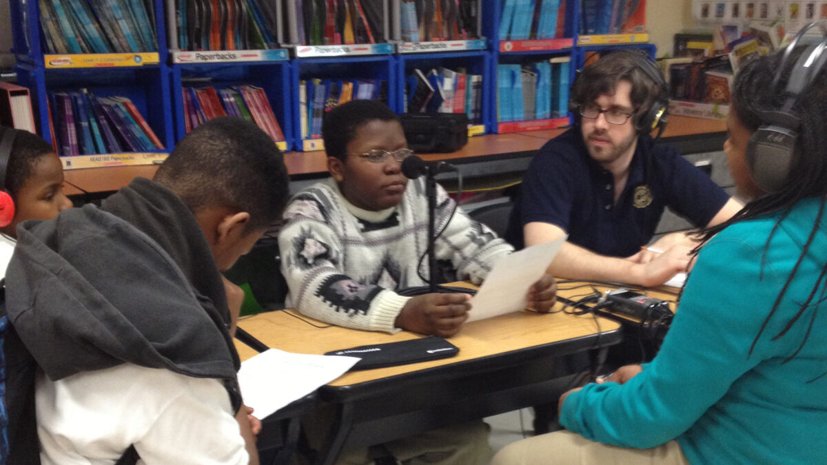A youth looking down at a piece of paper and two youth and an adult wearing headphones listening to a youth speaking into a microphone connected to a portable recorder and reading a piece of paper in a classroom