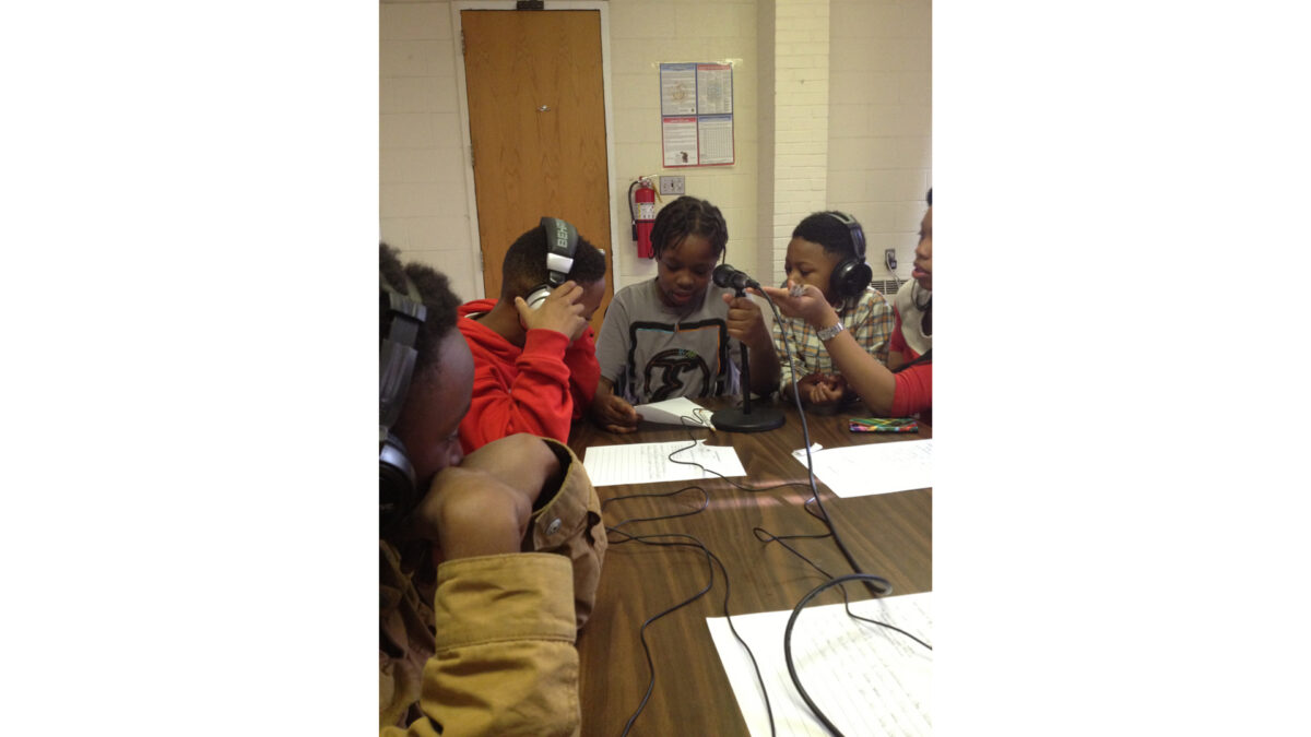 photo-3-(6)_wp Three youth wearing headphones listening to a youth reading a piece of paper into a microphone on a brown table