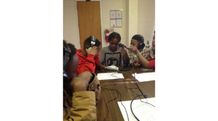 Three youth wearing headphones listening to a youth reading a piece of paper into a microphone on a brown table