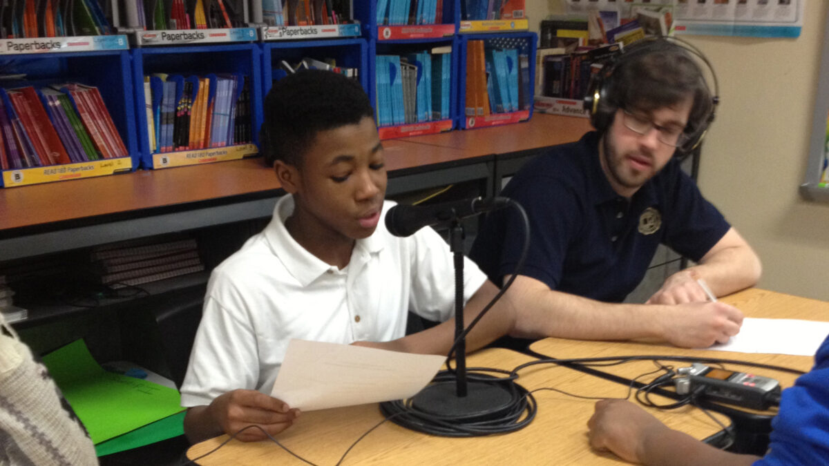 An adult wearing headphones listening to a youth reading a piece of paper into a microphone connected to a portable recorder in a classroom