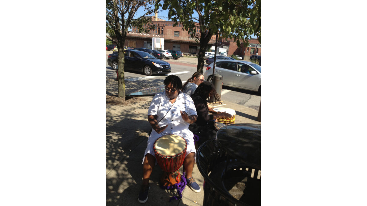 photo-4-(5)_wp Three adults playing djembes on a sidewalk next to a trash can in front of a red brick building