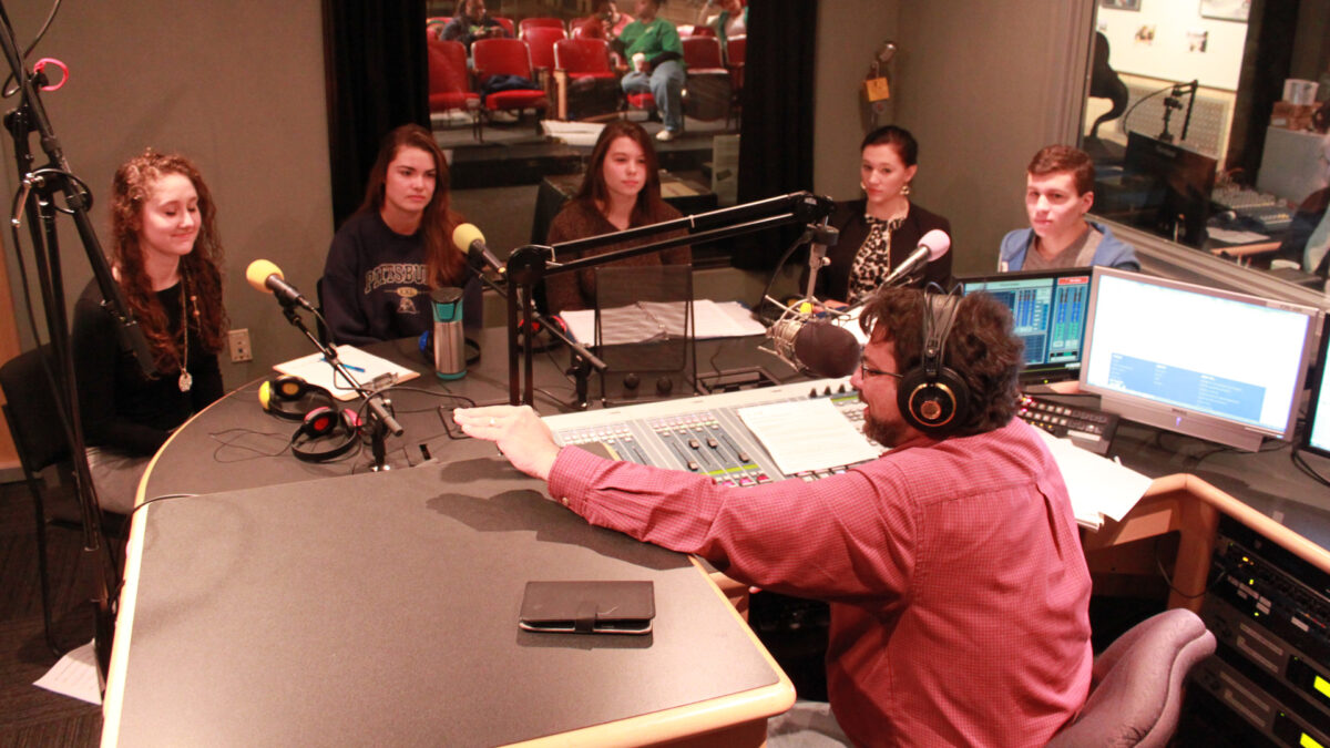 Four teens and one adult sitting behind microphones being interviewed by an adult wearing earphones and sitting behind a microphone and a mixing board gesturing with their hand in a radio studio