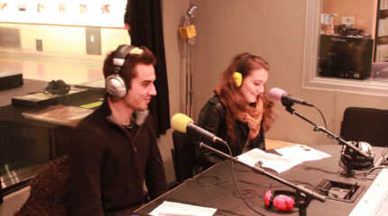 Two adults wearing headphones and speaking into microphones while looking at pieces of paper in a radio studio