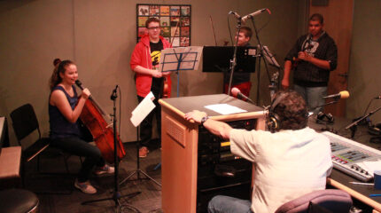 Two teens holding violins, one teen holding viola, and one teen holding a cello and speaking into a held microphone, all behind music stands being interviewed by and adult wearing headphones and sitting behind a mixing board in a radio studio