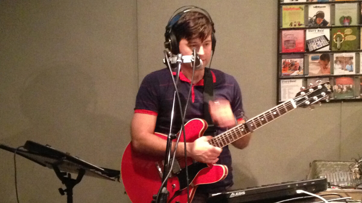 An adult wearing headphones holding an electric guitar and standing behind a microphone and a synthesizer in a radio studio