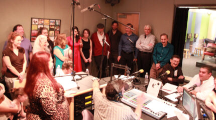 Sixteen adults standing around microphones and an adult wearing headphones sitting behind a microphone and a mixing board in a radio studio