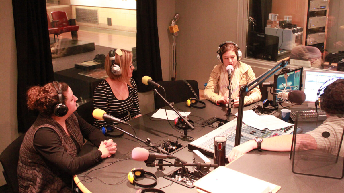 Three adults wearing headphones speaking into microphones being interviewed by an adult wearing headphones sitting behind a microphone and a mixer in a radio studio