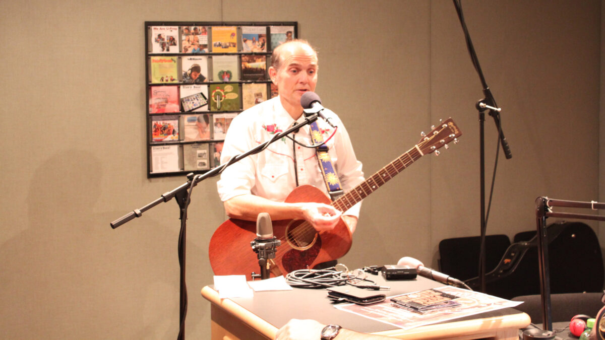 An adult holding an acoustic guitar and speaking into a microphone in a radio studio