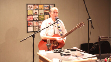 An adult holding an acoustic guitar and speaking into a microphone in a radio studio