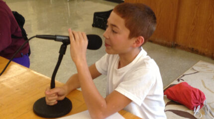 A youth holding a microphone with both hands while sitting at a classroom desk