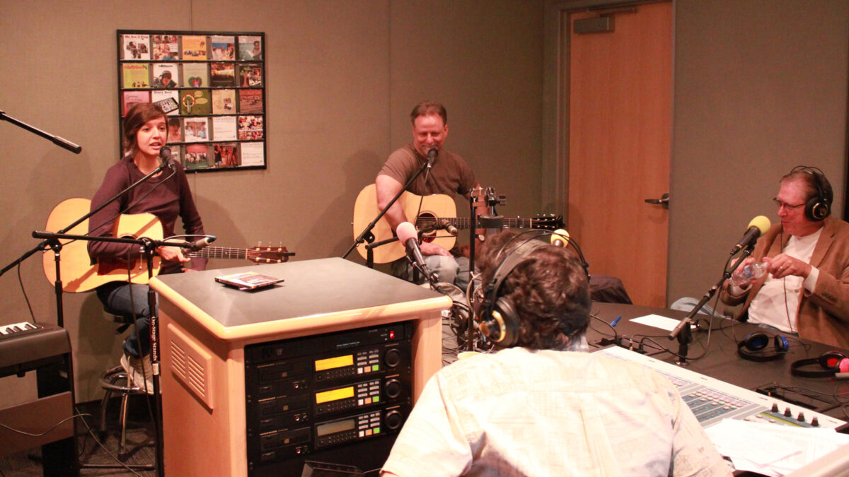 Two adults holding acoustic guitars and speaking into microphones and an adult wearing headphones sitting behind a microphone being interviewed by an adult wearing headphones sitting behind a mixer in a radio studio