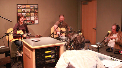 Two adults holding acoustic guitars and speaking into microphones and an adult wearing headphones sitting behind a microphone being interviewed by an adult wearing headphones sitting behind a mixer in a radio studio