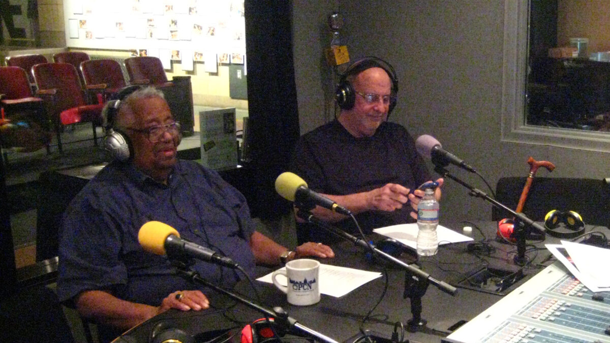 Two adults wearing headphones speaking into microphones in a radio studio