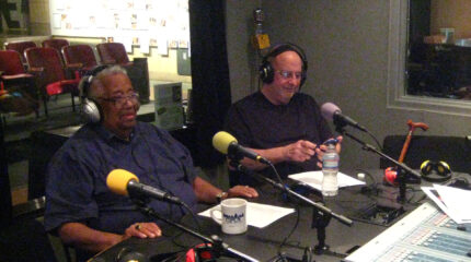 Two adults wearing headphones speaking into microphones in a radio studio