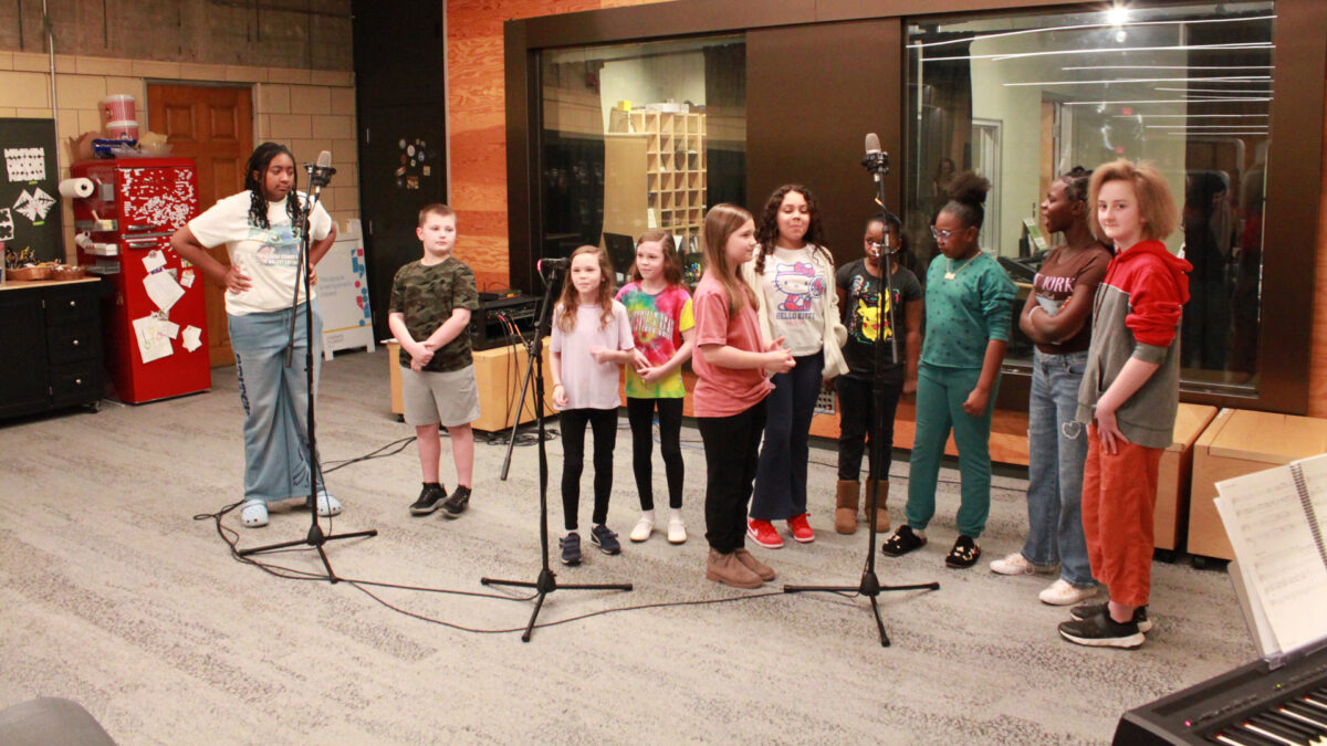 Ten youth singing into microphones in front of a radio studio