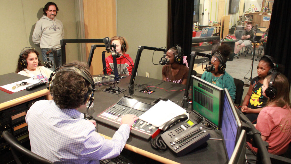 Six youth wearing headphones speaking into microphones being interviewed by an adult wearing headphones sitting behind a microphone and an audio mixer while another adult listens in the background in a radio studio