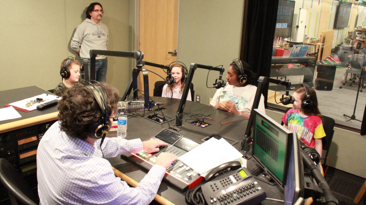Four youth wearing headphones speaking into microphones being interviewed by an adult wearing headphones sitting behind a microphone and an audio mixer while another adult listens in the background in a radio studio