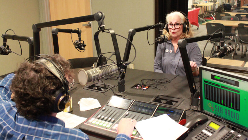 An adult wearing headphones speaking into a microphone being interviewed by an adult wearing headphones sitting behind a microphone and an audio mixer in a radio studio