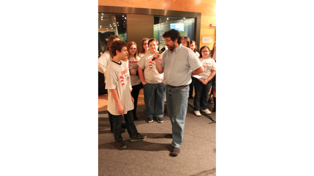 An adult holding and speaking into a microphone interviewing a youth as seven other youth watch, all standing in front of a radio studio