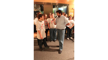 An adult holding and speaking into a microphone interviewing a youth as seven other youth watch, all standing in front of a radio studio