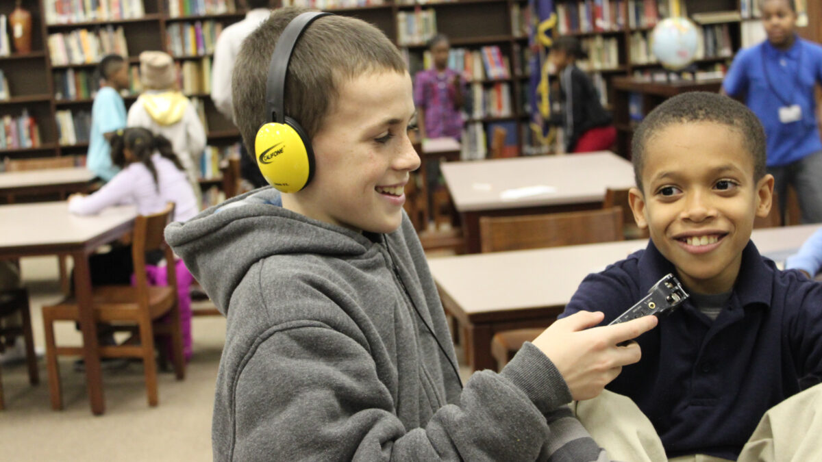A youth wearing headphones holding a portable recorder up to another youth smiling in a library