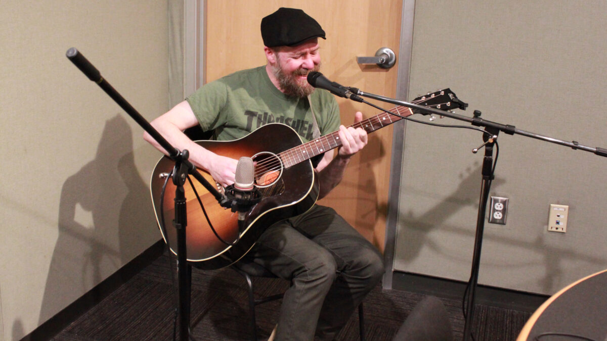 An adult playing an acoustic guitar and singing into a microphone in a radio studio