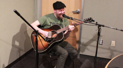 An adult playing an acoustic guitar and singing into a microphone in a radio studio