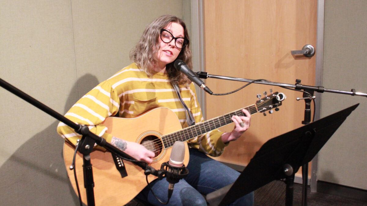 An adult sitting and singing into a microphone while playing an acoustic guitar in a radio studio