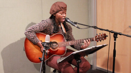 An adult playing acoustic guitar and singing into a microphone in a radio studio