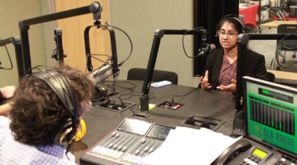 A teen wearing headphones speaking into a microphone being interviewed by an adult wearing headphones sitting behind a microphone and an audio mixer in a radio studio