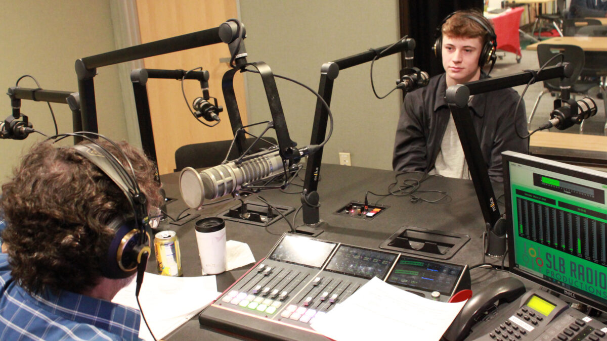 A teen wearing headphones sitting behind a microphone being interviewed by an adult wearing headphones sitting behind a microphone and an audio mixer in a radio studio