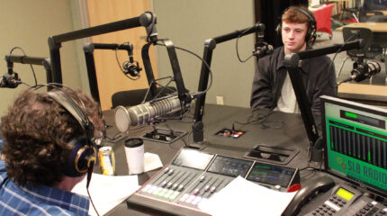 A teen wearing headphones sitting behind a microphone being interviewed by an adult wearing headphones sitting behind a microphone and an audio mixer in a radio studio
