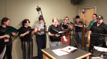 Eight adults singing into microphones while holding binders in a radio studio