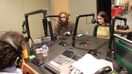 Two teens wearing headphones speaking into microphones being interviewed by an adult wearing headphones sitting behind a microphone and an audio mixer in a radio studio