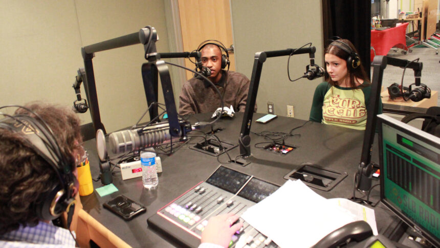 Two teens wearing headphones speaking into microphones being interviewed by an adult wearing headphones sitting behind a microphone and an audio mixer in a radio studio