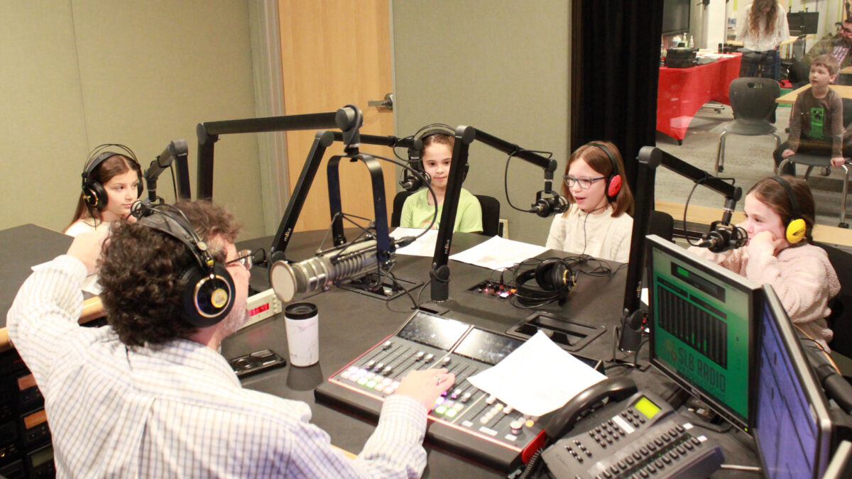 Four youth wearing headphones speaking into microphones being interviewed by an adult wearing headphones sitting behind a microphone and an audio mixer in a radio studio