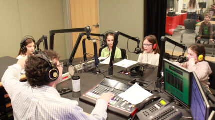 Four youth wearing headphones speaking into microphones being interviewed by an adult wearing headphones sitting behind a microphone and an audio mixer in a radio studio