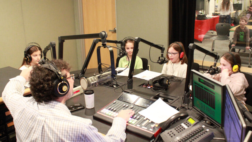 Four youth wearing headphones speaking into microphones being interviewed by an adult wearing headphones sitting behind a microphone and an audio mixer in a radio studio