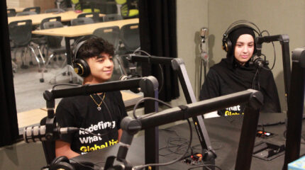 Two teens wearing headphones smiling and sitting behind microphones in a radio studio