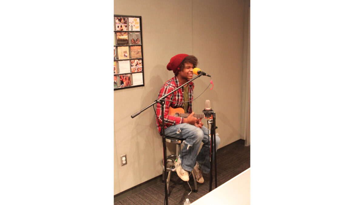 A teen sitting and smiling behind a microphone holding a ukulele in a radio studio