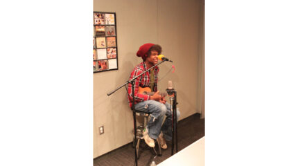 A teen sitting and smiling behind a microphone holding a ukulele in a radio studio