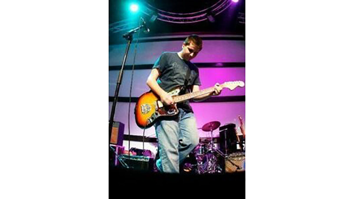A teen playing electric guitar standing behind a microphone and in front of a drum set on a stage