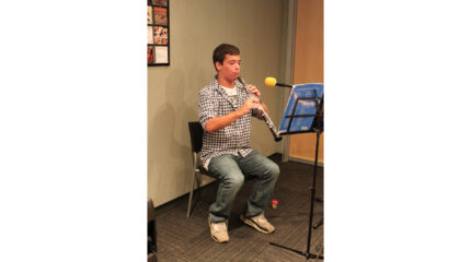 A teen sitting and playing oboe while looking at sheet music on a music stand in a radio studio