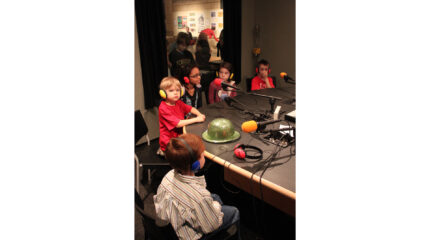 Five youth wearing headphones speaking into microphones in a radio studio