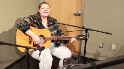 An adult sitting and playing an acoustic guitar while singing into a microphone in a radio studio