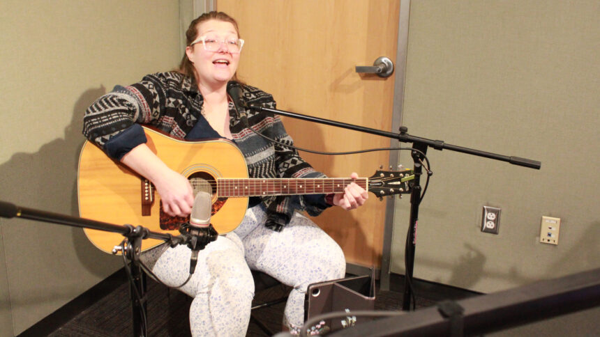 An adult sitting and playing an acoustic guitar while singing into a microphone in a radio studio